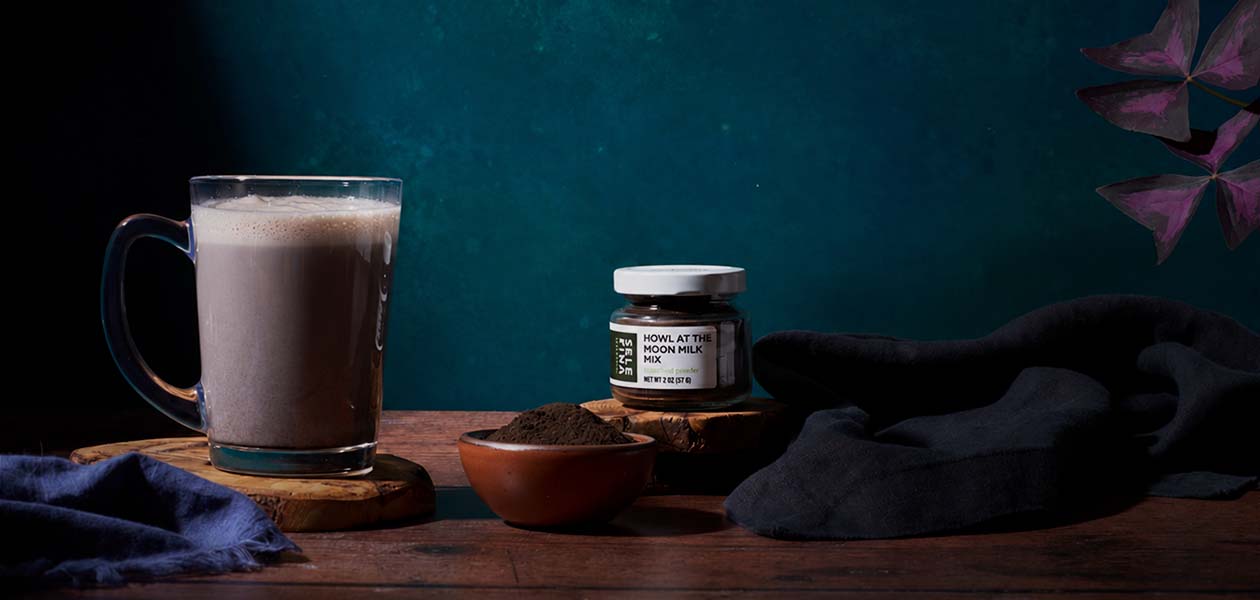 A mug of Howl at the Moon moon milk beside its jar and a bowl of the dark mix, against a deep teal wall